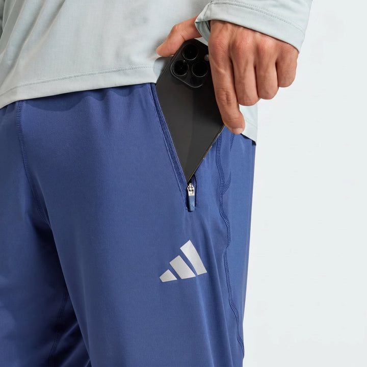 A close-up shot of a man wearing blue Adidas Adi365 Track Pants, placing a black smartphone into a zippered side pocket.