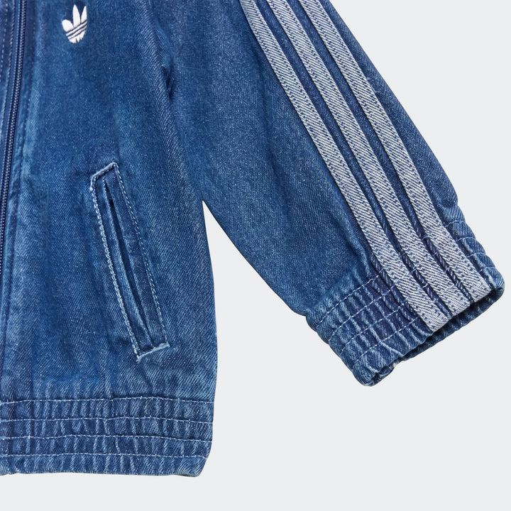 Close-up of a blue denim Adidas Firebird Track Top, showing the white trefoil logo, zipper, and three stripes on the sleeve.