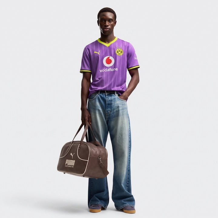 Man models a Borussia Dortmund 26/27 Third Jersey in purple and neon yellow, paired with wide-leg jeans and a brown duffel