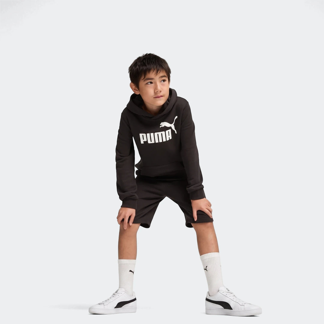 Young boy wearing black Puma Ess No. 1 Logo shorts and matching hoodie, white socks, and white sneakers, posing in a studio.