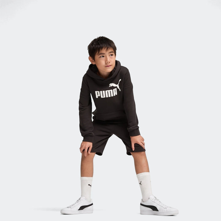 Young boy wearing black Puma Ess No. 1 Logo shorts and matching hoodie, white socks, and white sneakers, posing in a studio.