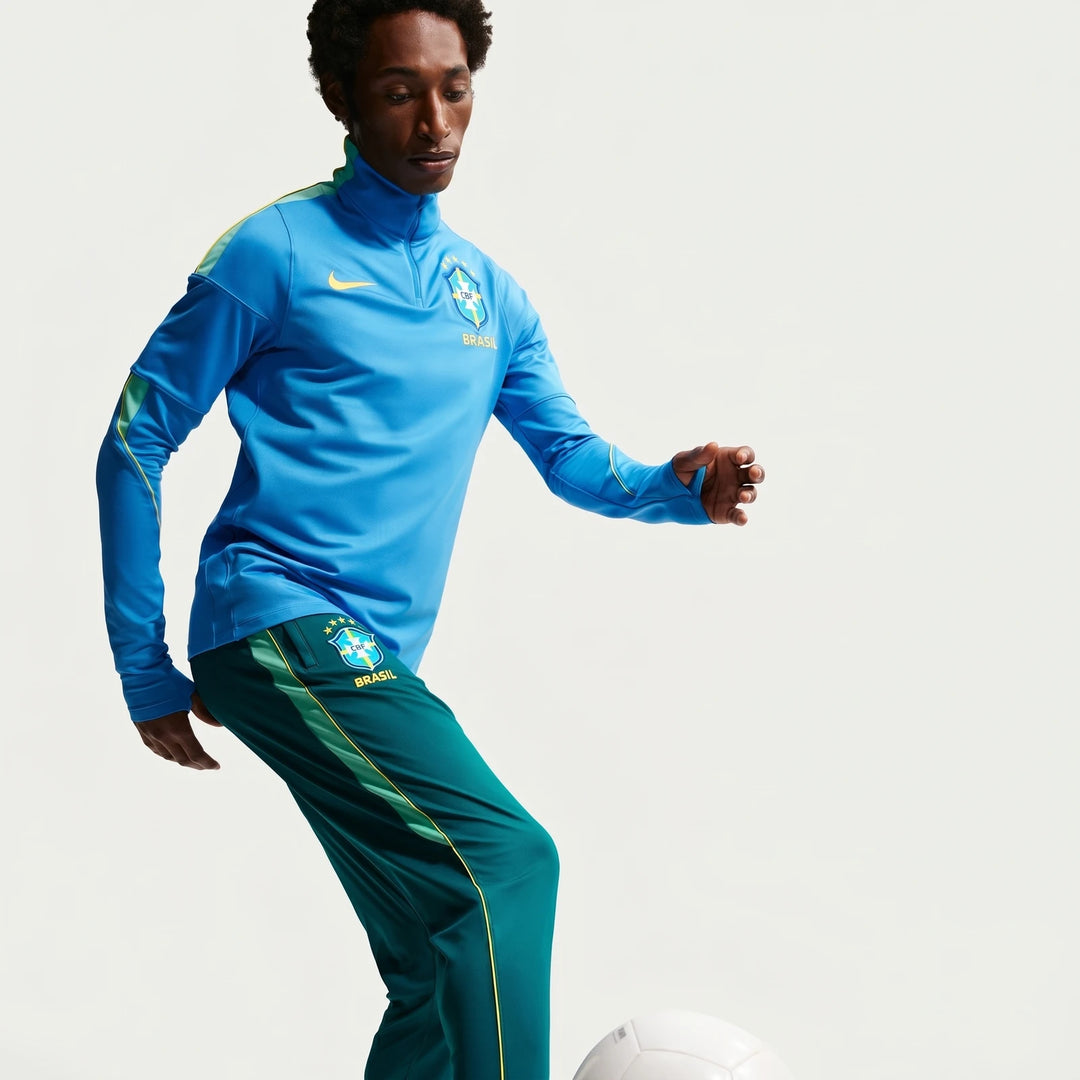Male model in a blue Nike Brazil soccer quarter-zip top and green track pants playing with a white soccer ball.