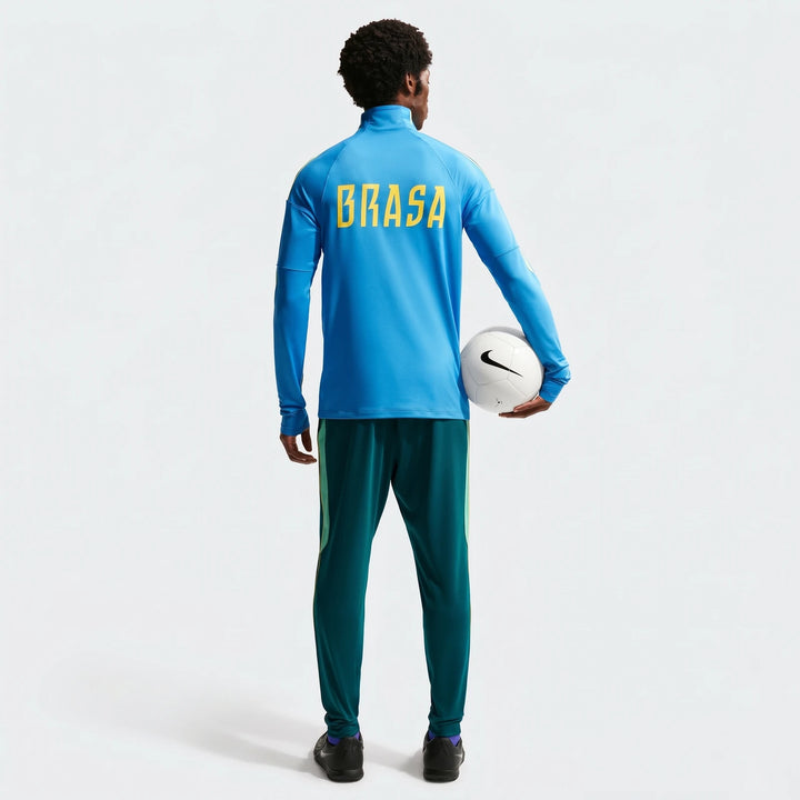 A man from behind wearing a blue Nike Dri-Fit long-sleeve top with "BRASA" and green soccer track pants, holding a white