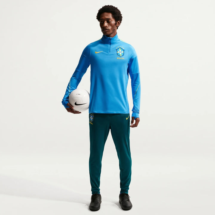 Brazil Academy Pro Green Men's Nike Dri-Fit Soccer Track Pants worn by a model in a blue top, holding a white soccer ball.