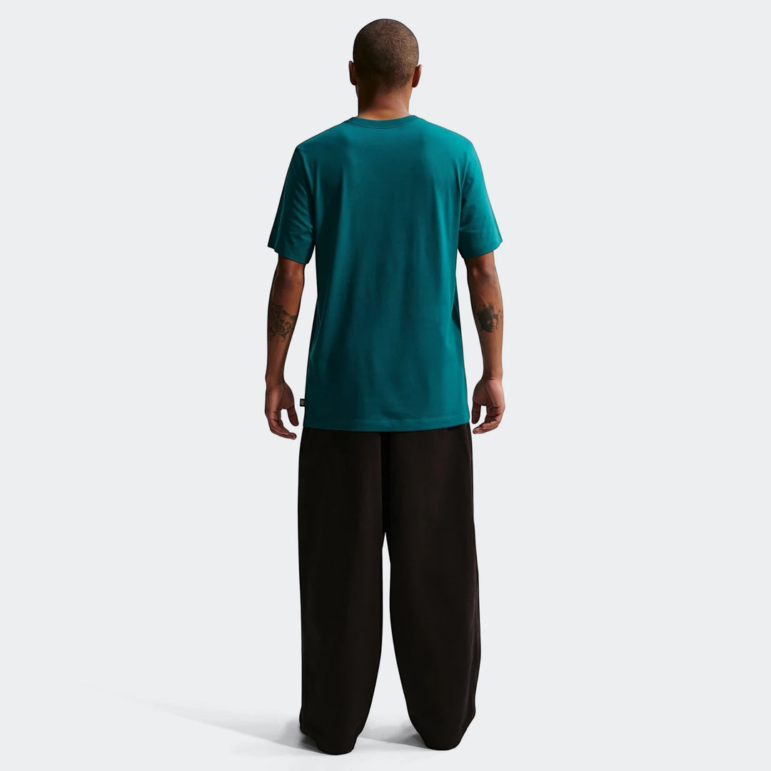 Rear view of a man wearing a Brazil Green Men's Nike Soccer T-Shirt and black wide-leg pants on a white background.