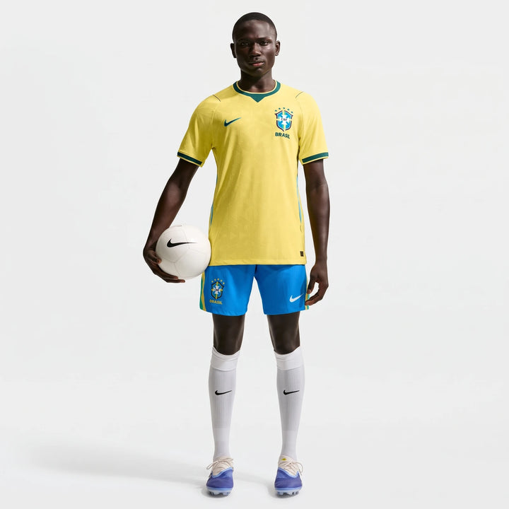 Male model in yellow Brazil National Team 2026 Match Home Nike Aero-Fit soccer jersey and blue shorts, holding a white