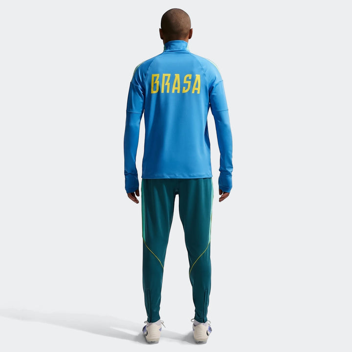 Back view of a male model wearing a Brazil Strike blue Nike Dri-Fit long-sleeve soccer drill top and dark teal track pants.