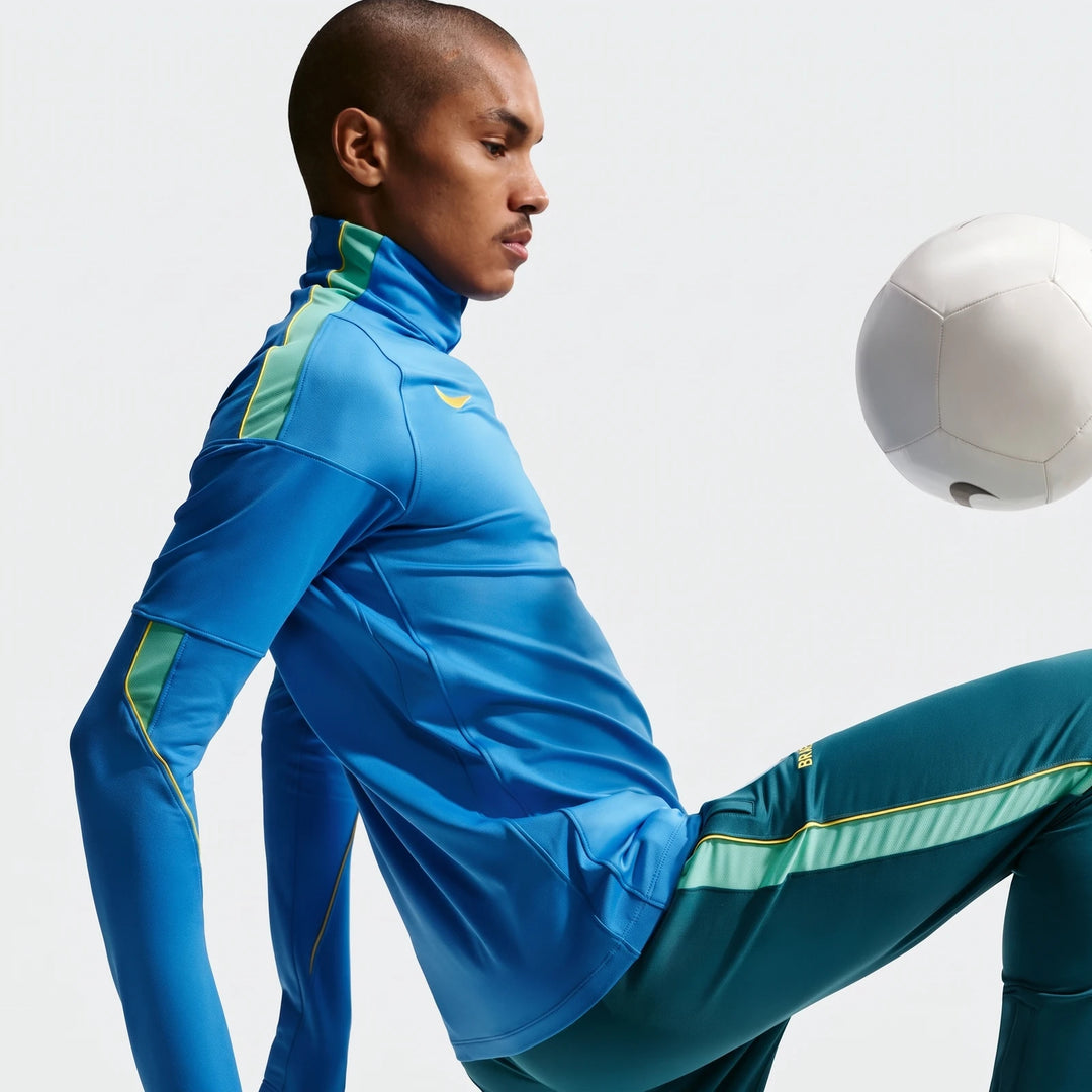 Male model wearing a blue and teal Nike Dri-FIT soccer drill top and matching pants, in a dynamic pose with a white soccer