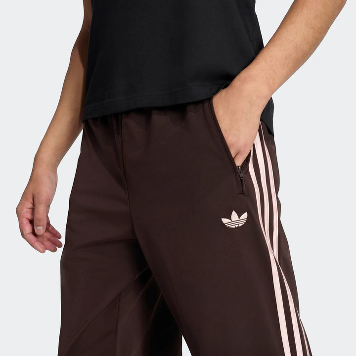 Close-up of brown Adidas Firebird Track Pants with light pink triple stripes and Trefoil logo, worn by a model with hand in