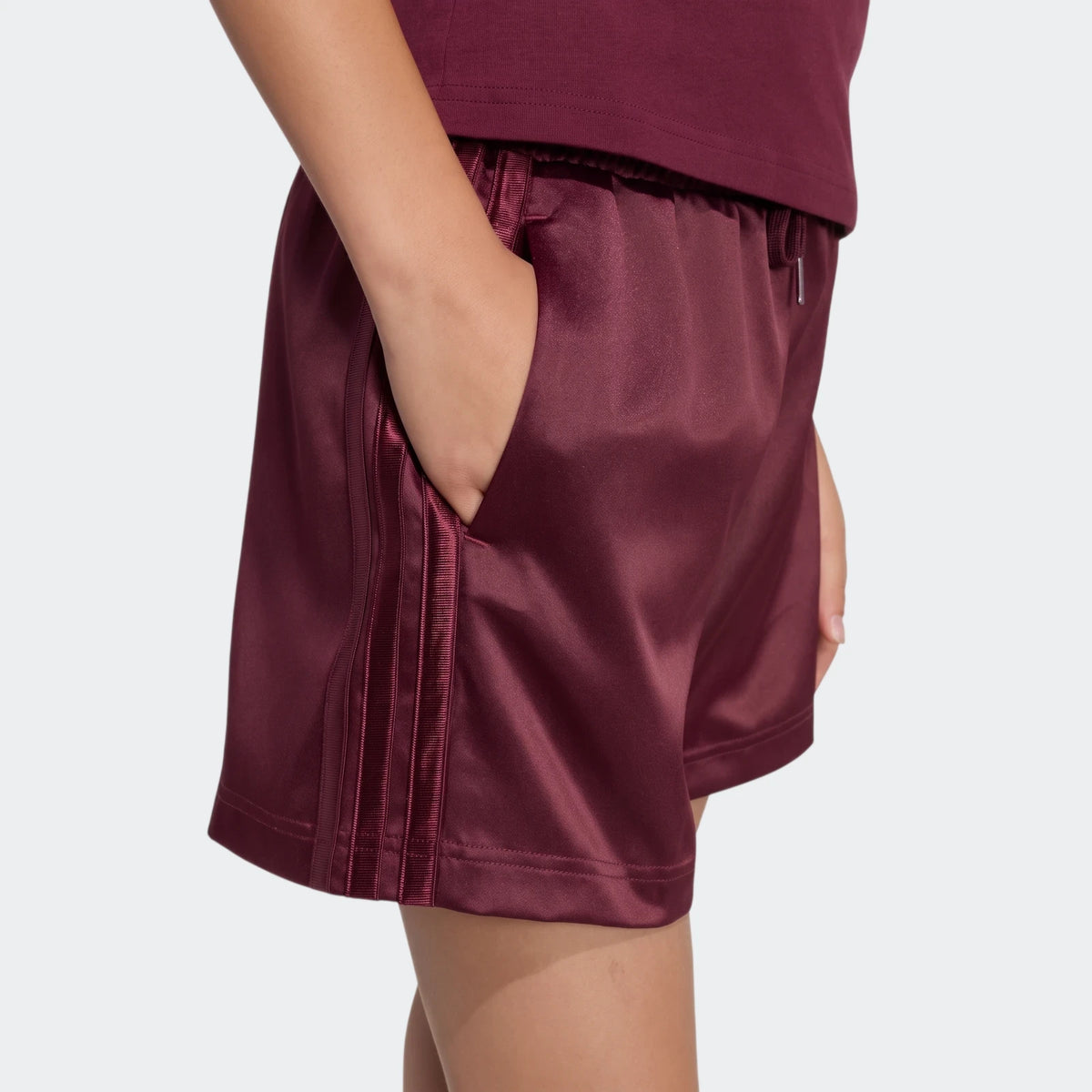 Side view of burgundy satin shorts with three vertical stripes, worn by a person with their hand in the pocket.