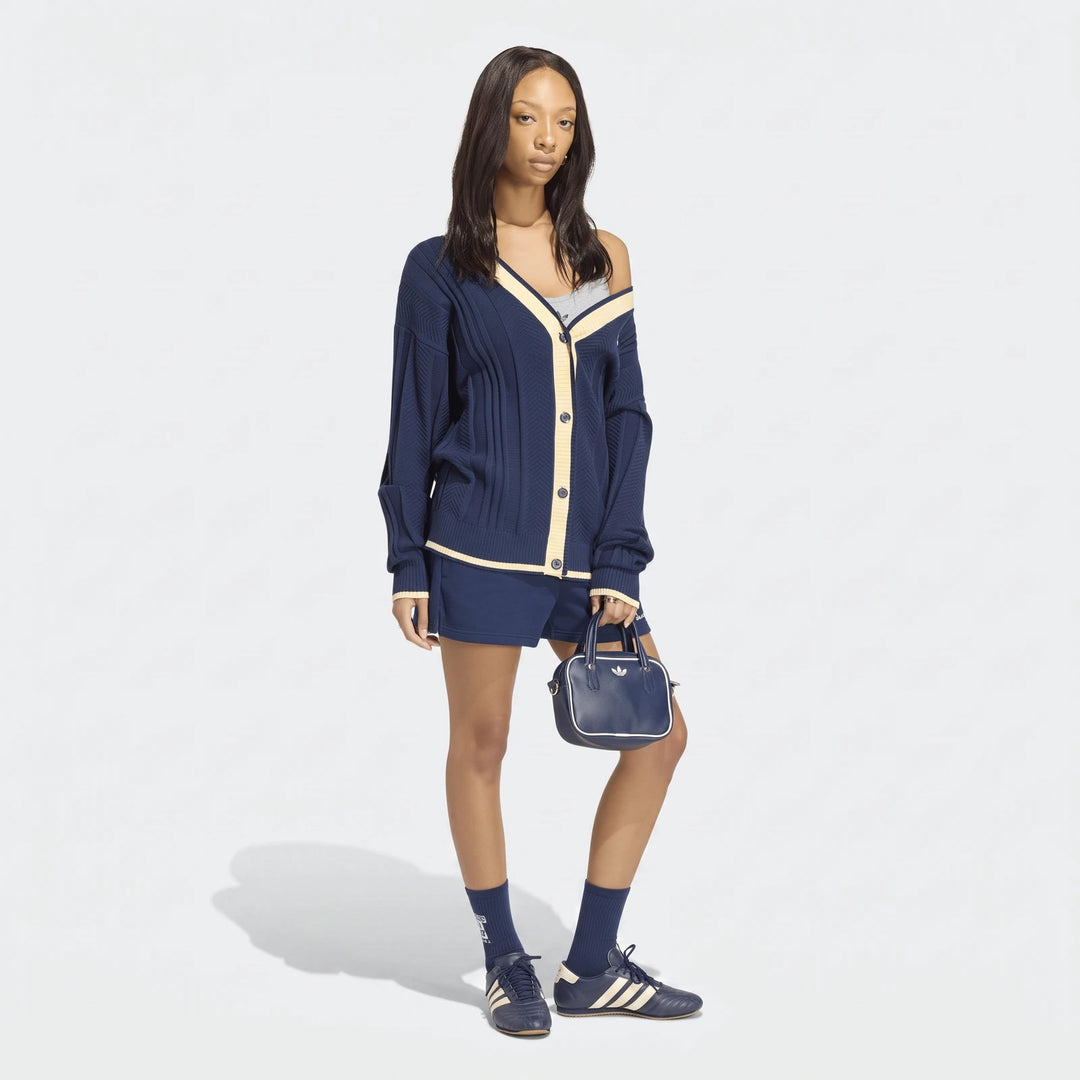 Female model wearing a navy blue cable knit cardigan, matching shorts, and sneakers, holding a small Adidas bag.
