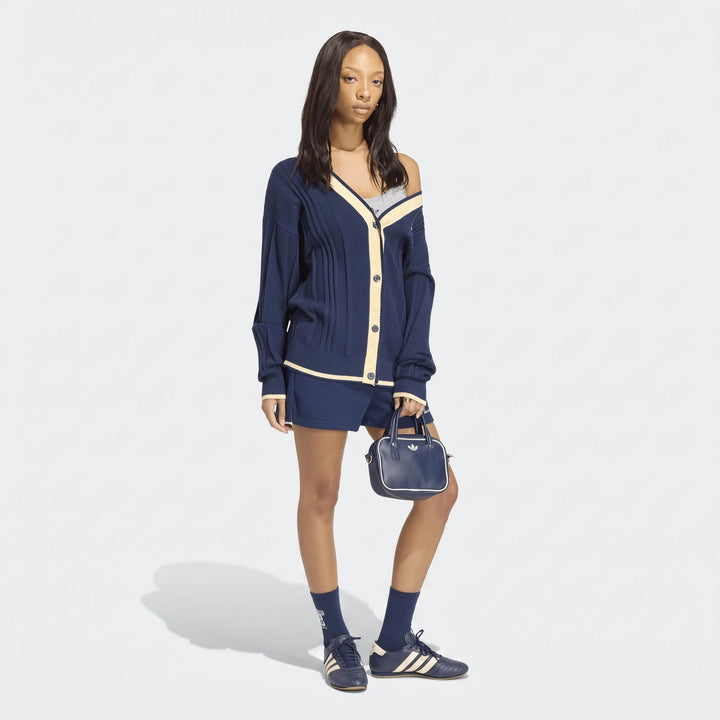 Female model wearing a navy blue cable knit cardigan, matching shorts, and sneakers, holding a small Adidas bag.