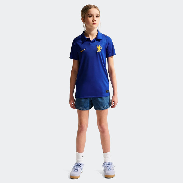 Young girl wearing Chelsea FC 2026/27 Stadium Home Blue Big Kids' Nike Dri-Fit Soccer Replica Jersey and blue denim shorts.