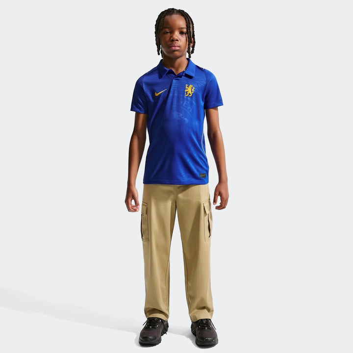 Young boy wearing a blue Nike Chelsea FC 2026/27 Stadium Home soccer replica jersey with khaki cargo pants.