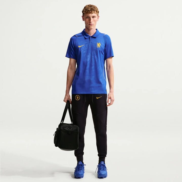 Male model wears Chelsea FC 2026/27 Stadium Home Nike Dri-Fit Soccer Replica Jersey, black track pants, and blue sneakers,
