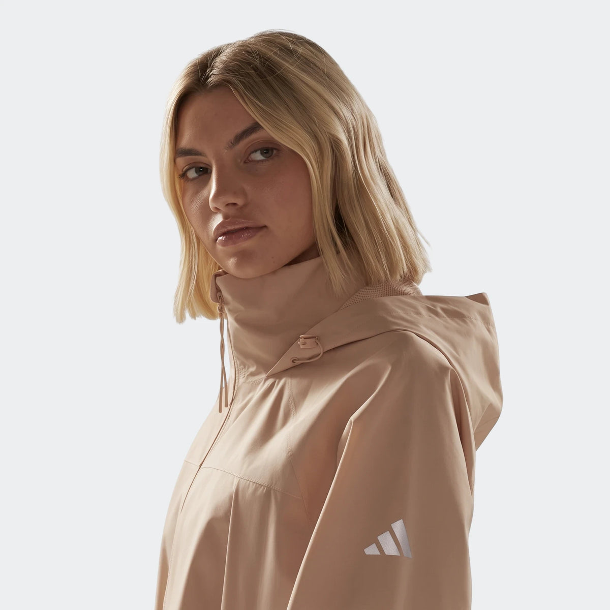 Close-up of a woman wearing a light brown Adidas City Rain Parka with a hood and high collar.