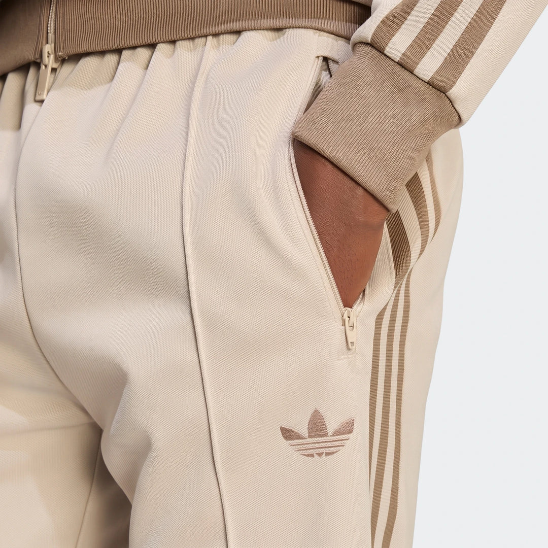 Close-up of classic beige Adidas track pants worn by a person, featuring zippered pockets and signature brown three-stripe