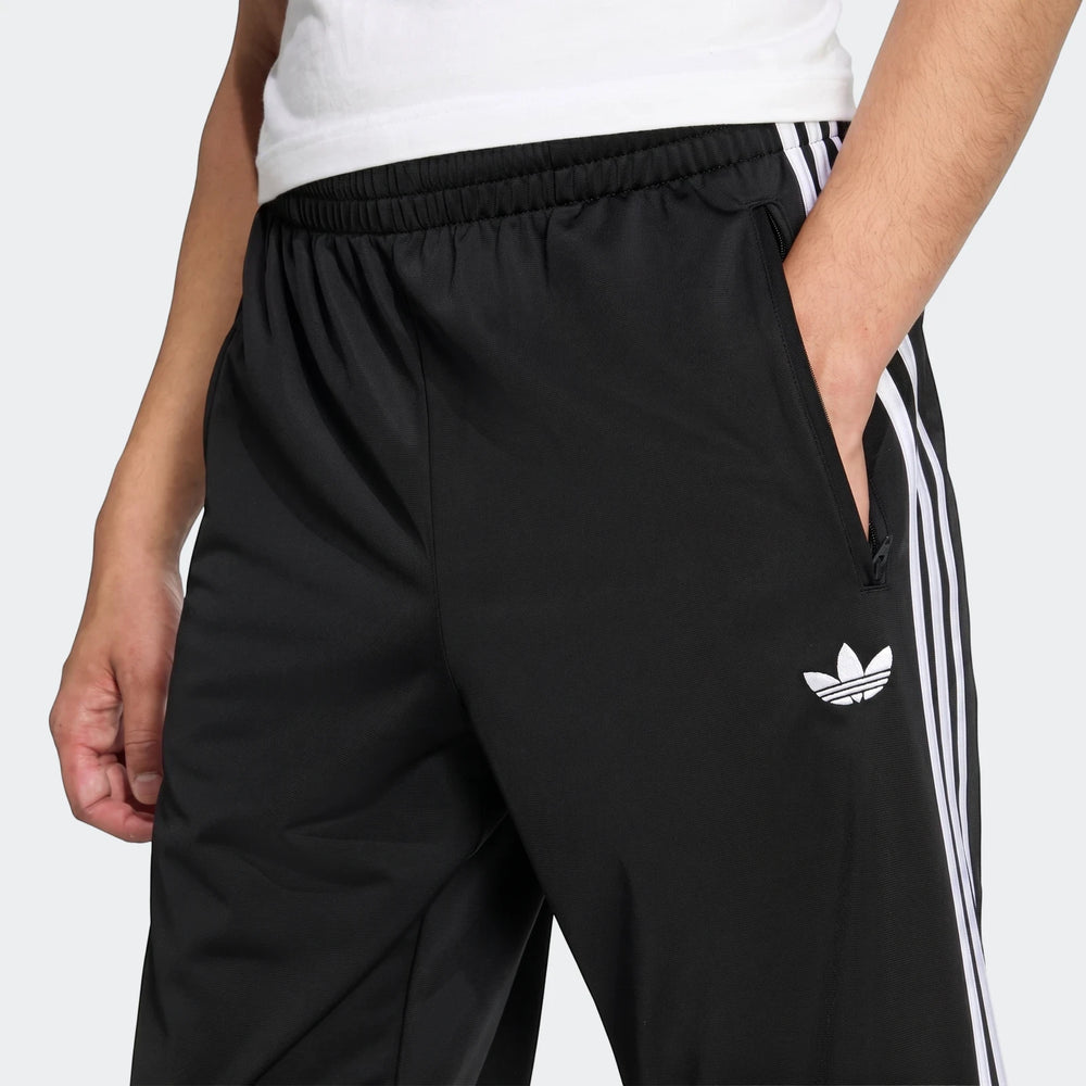 Close-up of black Adidas Firebird Track Pants with white three stripes and trefoil logo, worn by a man with his hand in the