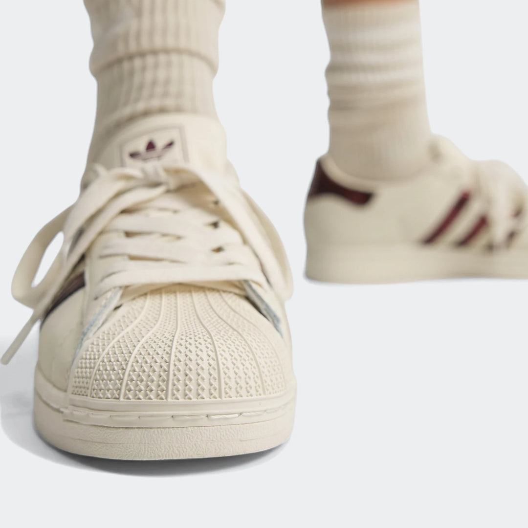 Close-up of a person wearing creamy white Adidas Superstar II shoes with dark red stripes and iconic shell toes. The shoes