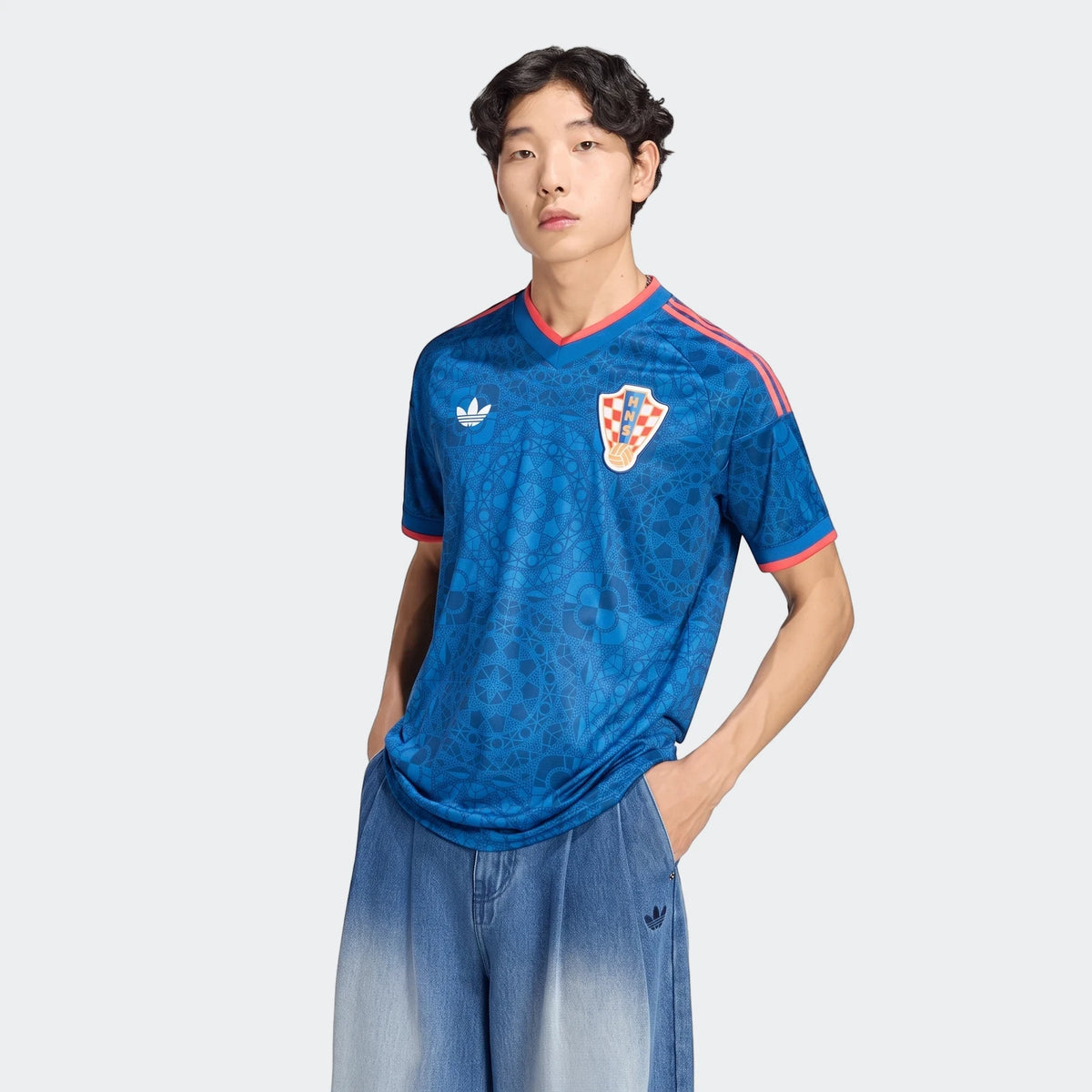 Male model wearing a blue patterned Adidas Croatia 26 Away Jersey with red trim and light wash jeans, posed mid-thigh up.