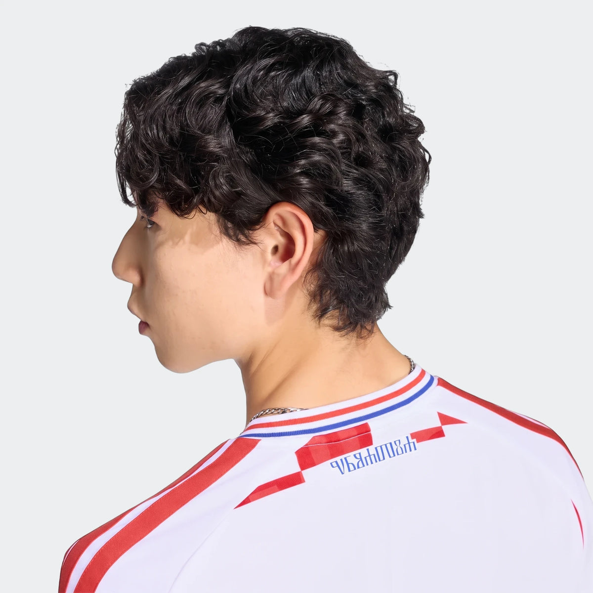 A young man from the back and side, wearing a white Croatia 26 Home Jersey with red stripes and blue details.