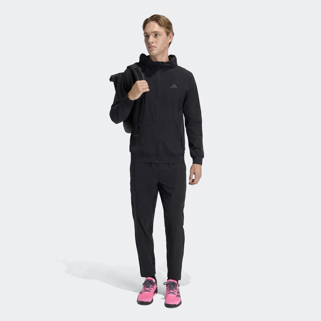 Male model in a black Adidas D4T Hybrid Full-Zip Hoodie, black track pants, pink sneakers, and a black backpack, standing