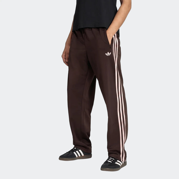 Dark brown Adidas Firebird Track Pants with light pink stripes and trefoil logo, worn by a person, paired with black