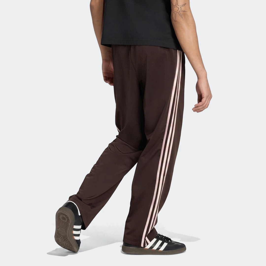A person from the waist down wearing dark brown Firebird Track Pants with white stripes and black sneakers, walking away.