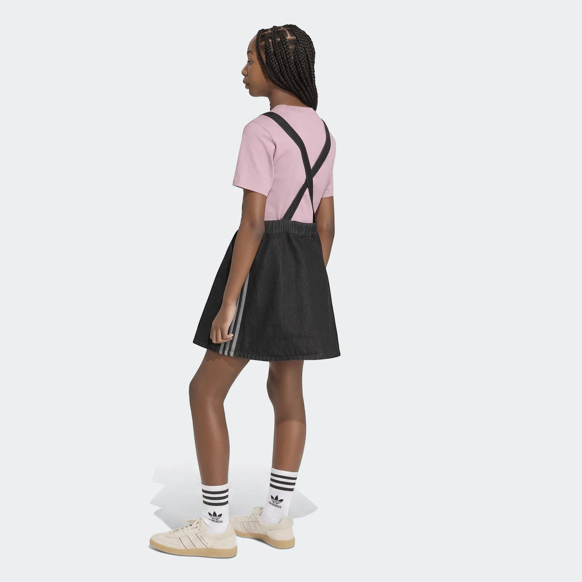 Young girl models a dark denim dungarees dress with a pink t-shirt, white striped socks, and beige sneakers.