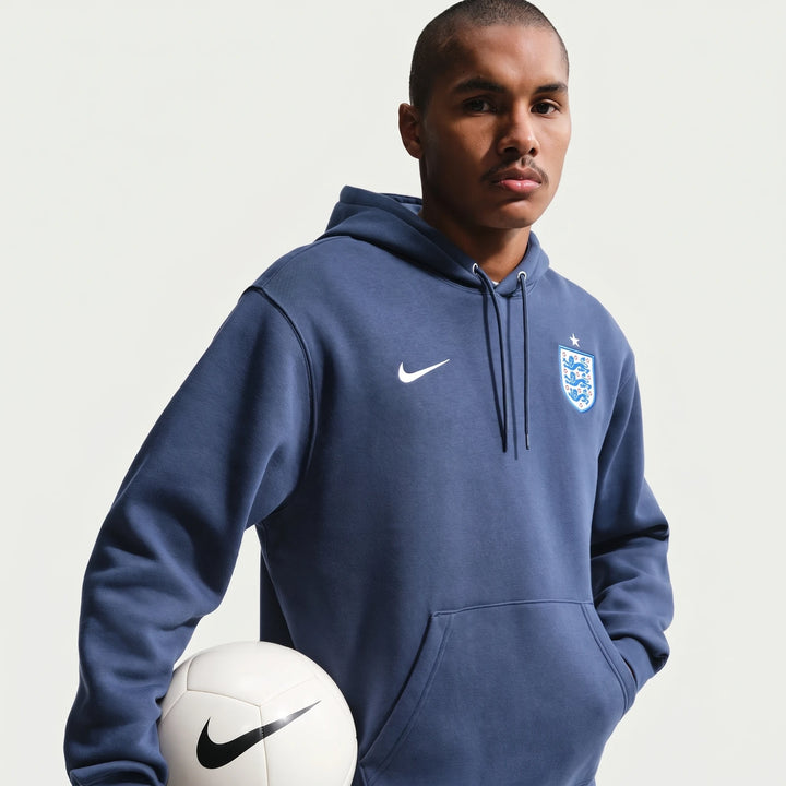 Men's England Club blue Nike soccer pullover hoodie worn by a model, holding a white soccer ball.