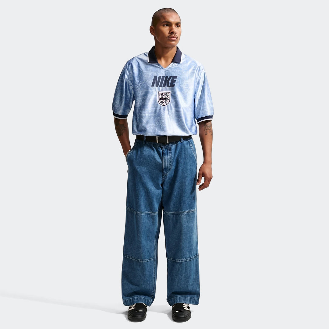 A male model wears a light blue Nike England Energy men's Dri-Fit short-sleeve soccer top with wide-leg blue jeans and black