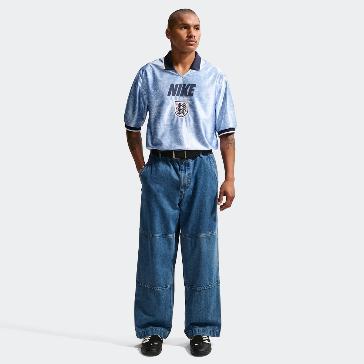 A male model wears a light blue Nike England Energy men's Dri-Fit short-sleeve soccer top with wide-leg blue jeans and black