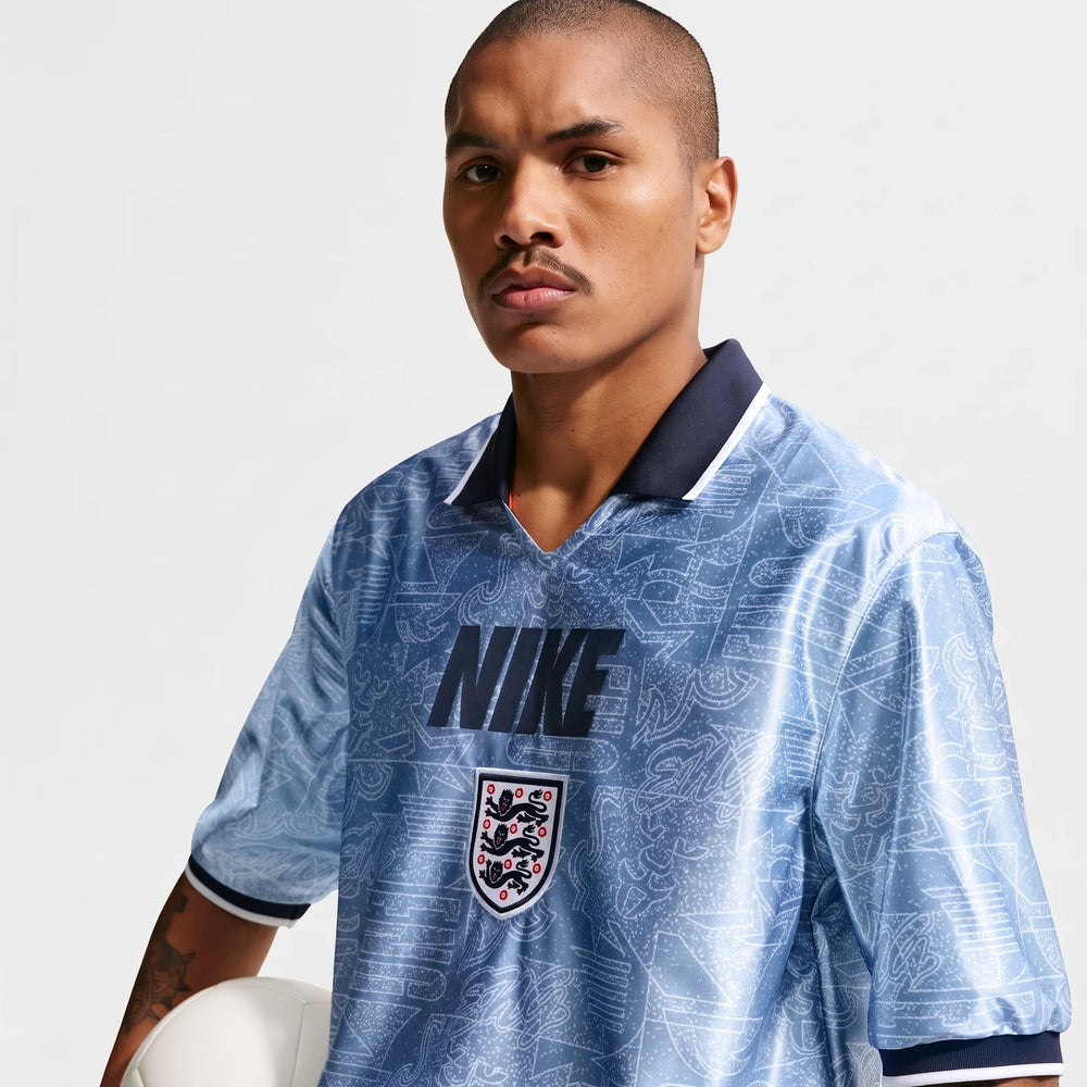 Male model wearing a light blue patterned Nike Dri-Fit short-sleeve soccer top with an England crest, holding a white soccer