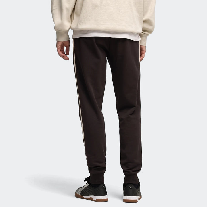 Rear view of men's Ess Elevated dark brown sweatpants with light side stripes, worn with sneakers.