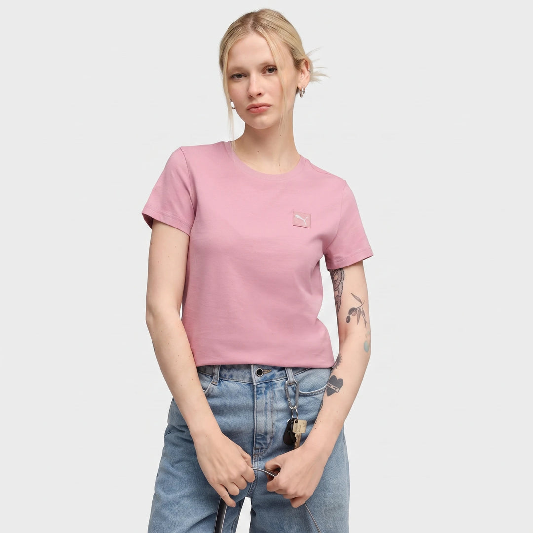 A woman models the Ess Elevated Tee W in pink, featuring a small logo. She wears blue jeans and has blonde hair pulled back.