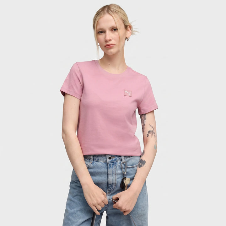 A woman models the Ess Elevated Tee W in pink, featuring a small logo. She wears blue jeans and has blonde hair pulled back.