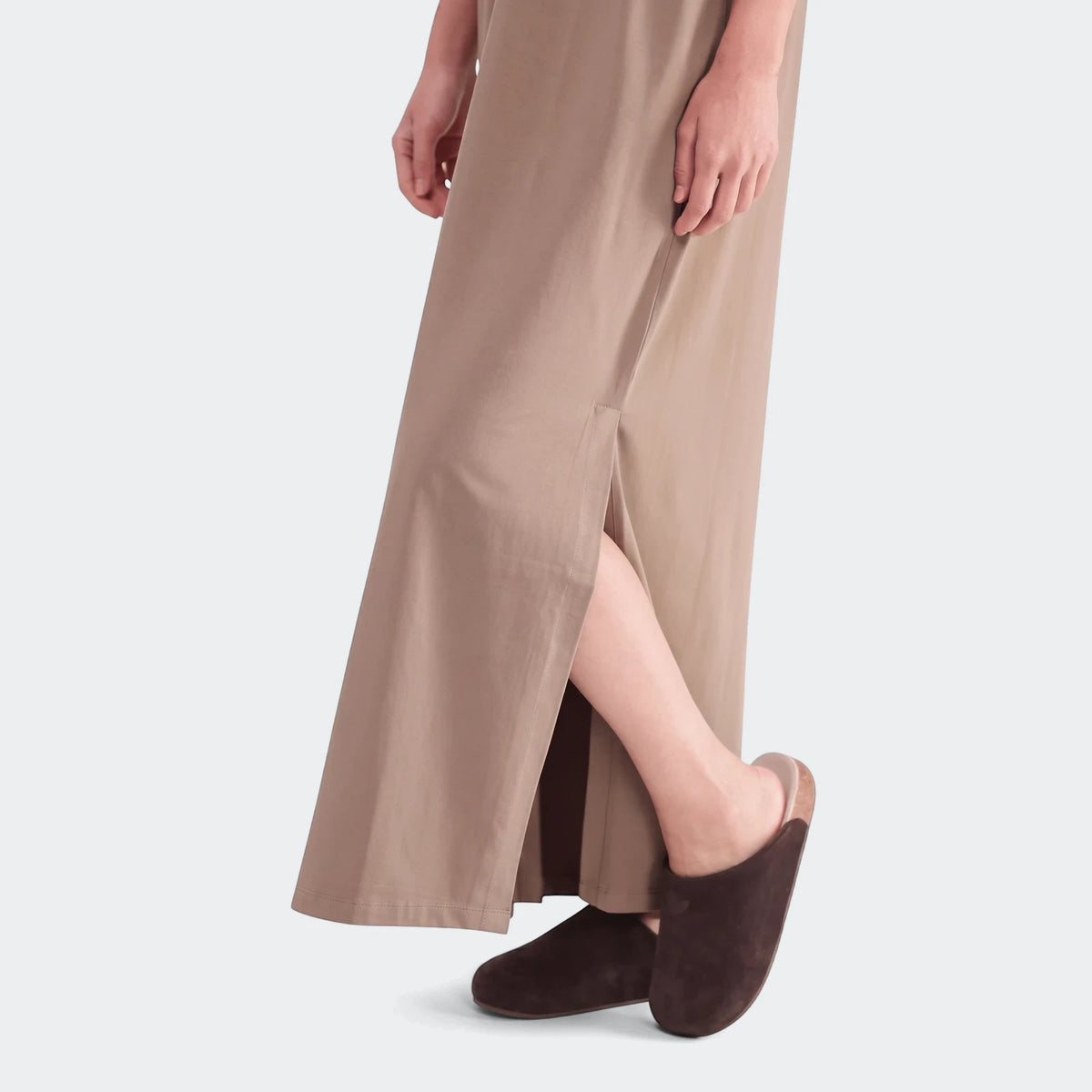 Model wearing a loose-fitting long beige essential dress with a side slit, paired with dark brown slippers.