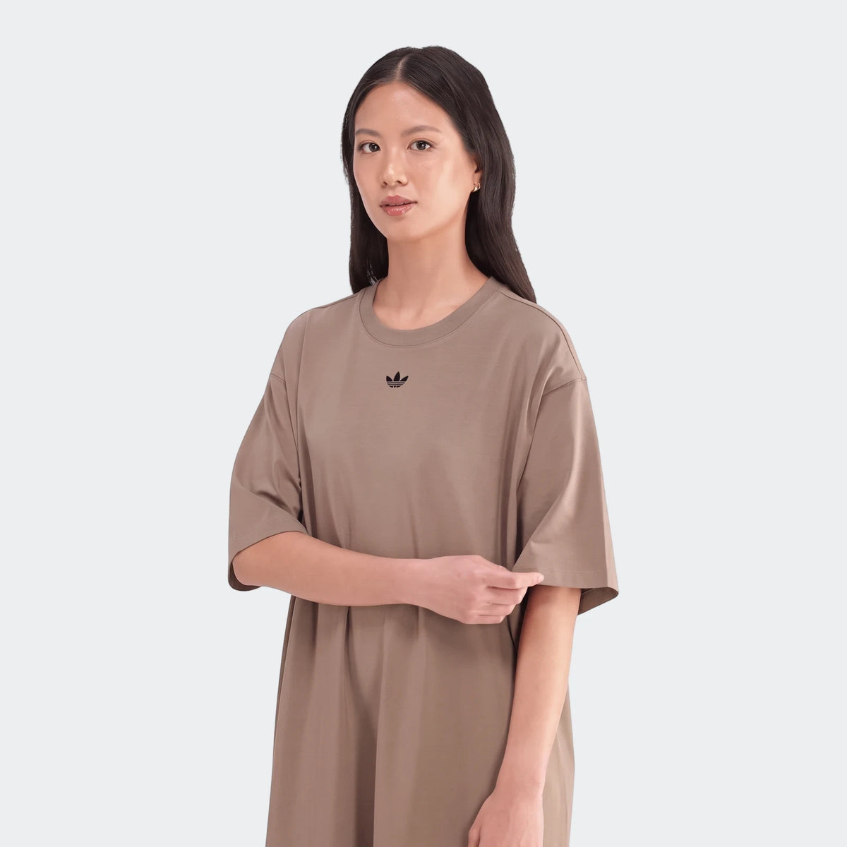Woman models a loose-fitting, short-sleeve Essential Beige Dress with a black Adidas Trefoil logo. The casual dress features