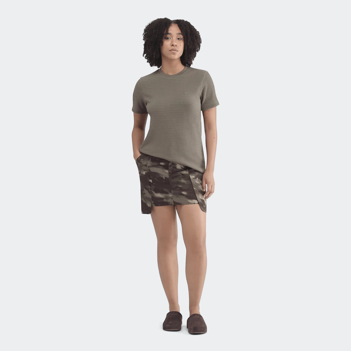 An essential olive green waffle knit t-shirt worn by a woman, paired with a camouflage mini skirt and dark brown shoes.
