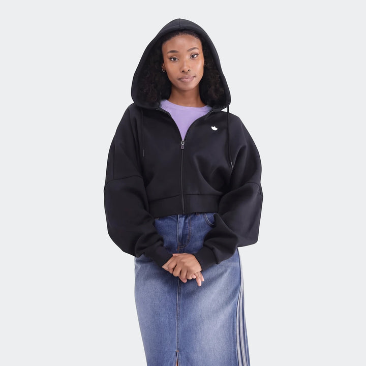 A woman models a black Essentials full-zip cropped hoodie with a white logo, paired with a blue denim midi skirt. Full body