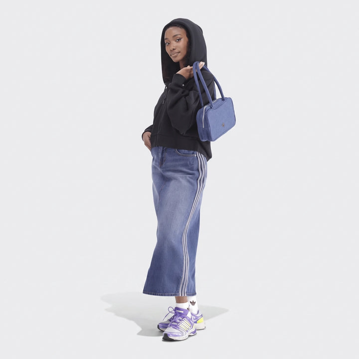 A female model wears a black Essentials Full-Zip Hoodie, a denim midi skirt, purple sneakers, and carries a blue denim