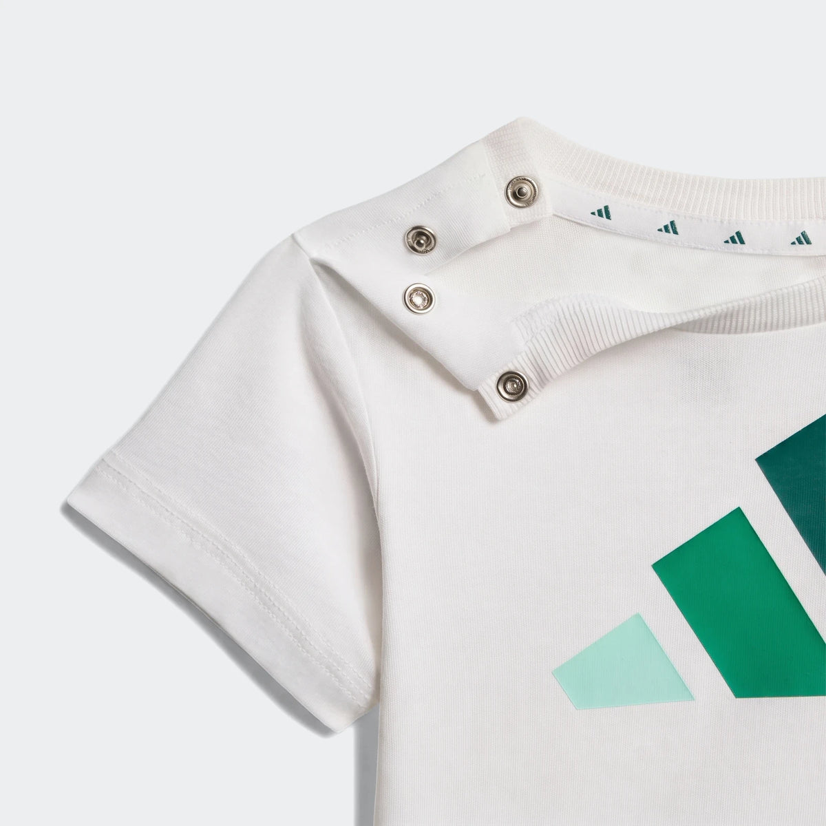 Close-up of a white short-sleeve Essentials Tee Set Kids t-shirt with green Adidas logo and silver shoulder snaps.