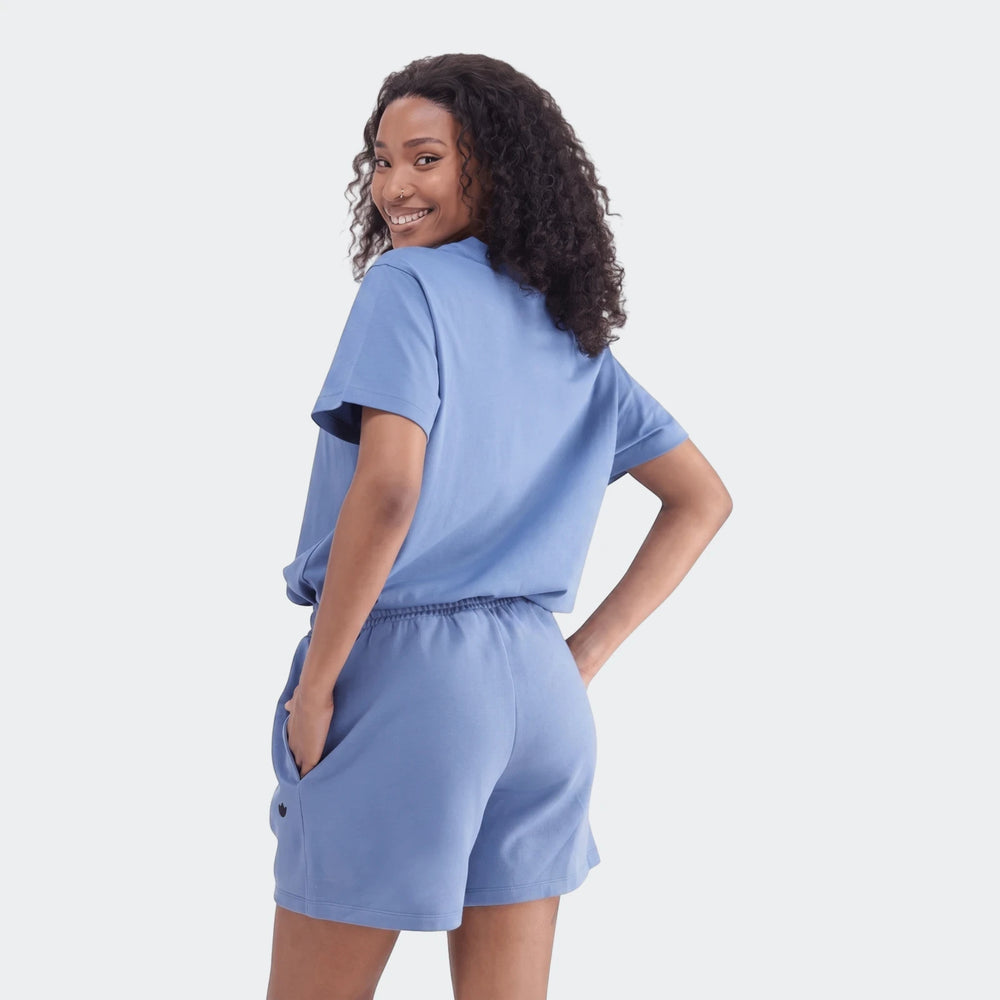 Young woman smiling, wearing a blue Essentials Trefoil T-Shirt and matching blue shorts. She has dark curly hair and a nose