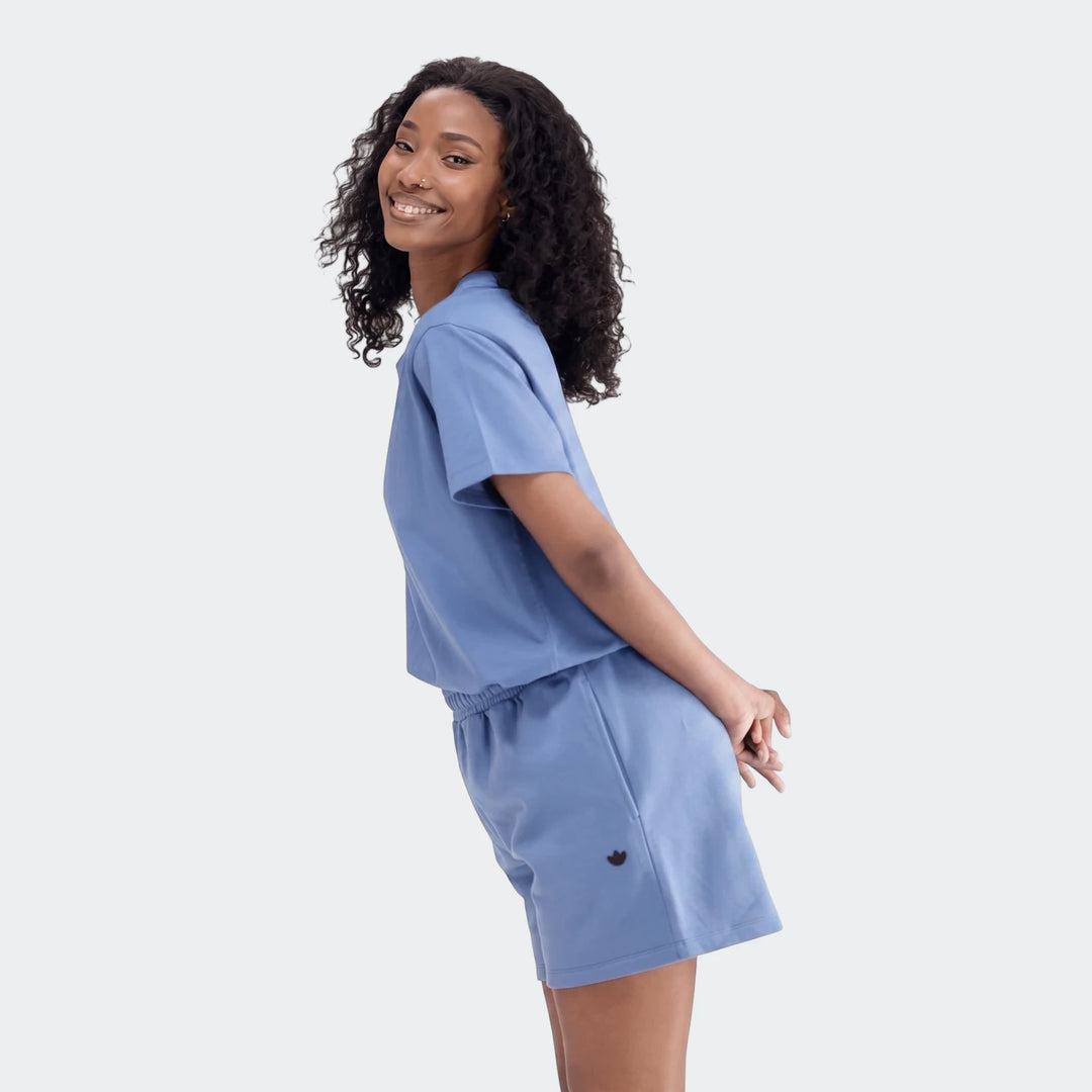 Smiling young woman wearing a blue Essentials Trefoil T-shirt and matching blue shorts, posing on a plain background.