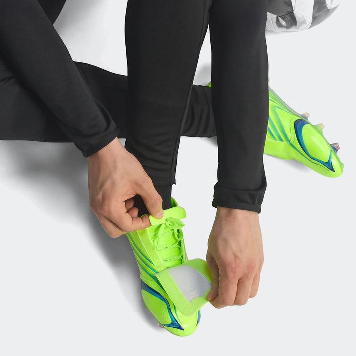 A person in black athletic pants kneels to tie the laces of bright neon green and blue F50 Elite FG Tunit football boots.