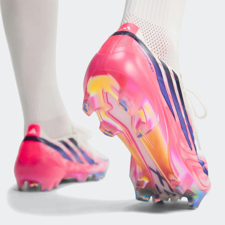 Pink and iridescent Adidas F50 Hyperfast Elite firm ground football boots worn with white socks, showing cleats.