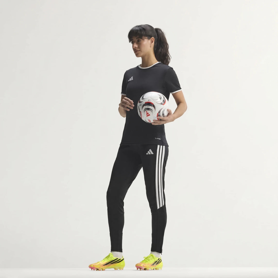 Woman in black Adidas training kit, holding a soccer ball and wearing yellow F50 Hyperfast Elite Firm Ground Football Boots.