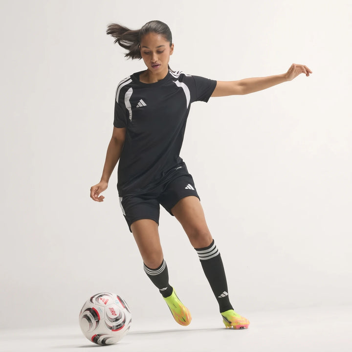 Female soccer player in black Adidas kit and F50 Hyperfast Elite Laceless Firm Ground Boots kicking a soccer ball.