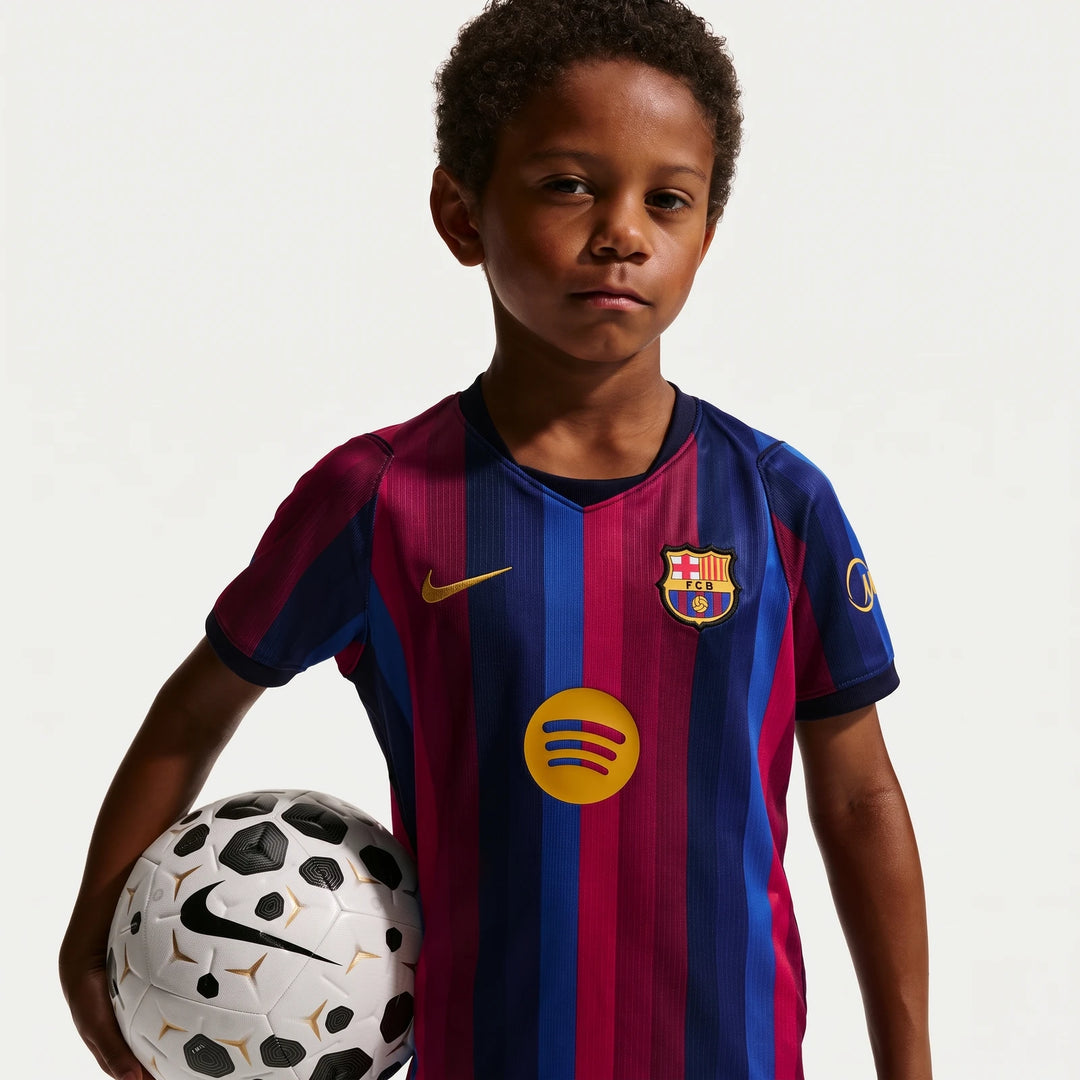A young boy wearing the blue and red striped FC Barcelona 2026 Stadium Home Nike Soccer Replica Kit, holding a white and