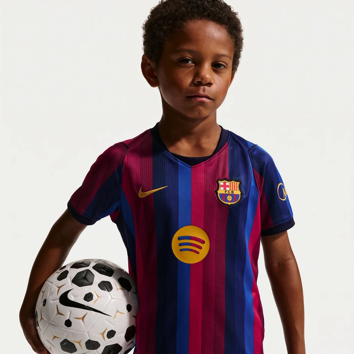 A young boy wearing the blue and red striped FC Barcelona 2026 Stadium Home Nike Soccer Replica Kit, holding a white and